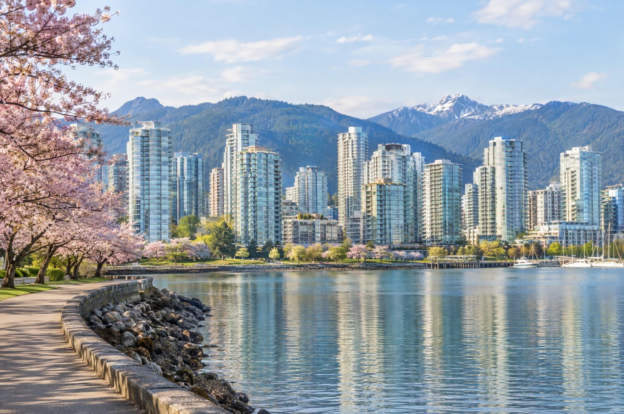Downtown waterfront towers seen across calm water from a seawall path, mountains behind